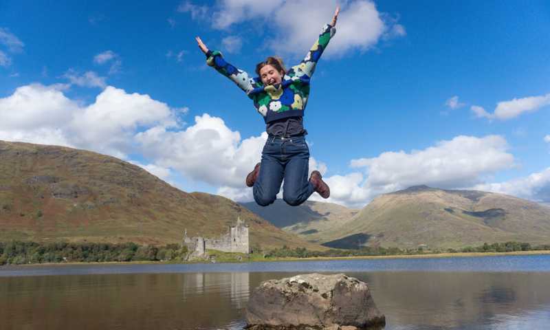 Young woman jumping with sunny views of Kilchurn Castle across the shore