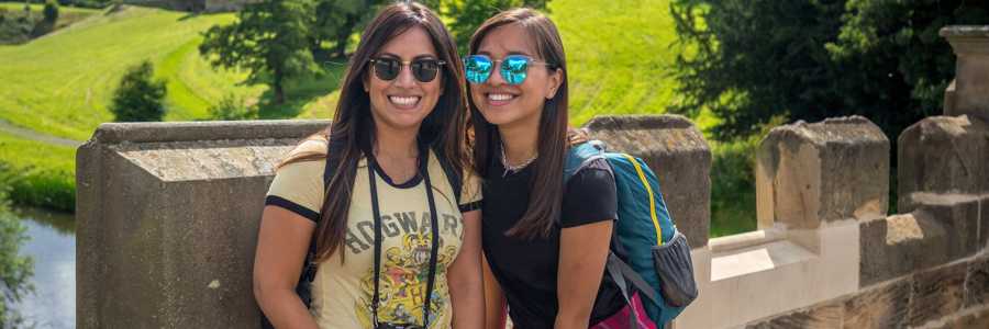 Two young women posing on a stone bridge with Alnwick Castle’s grand towers in the sunny background