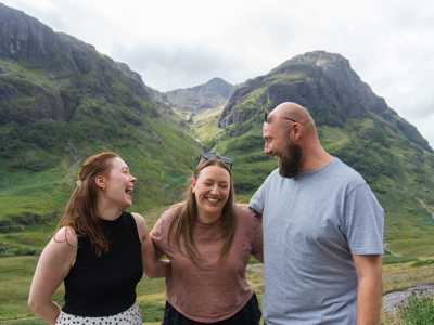 Three friends laughing together in Glen Coe, surrounded by rugged peaks and lush summer greenery
