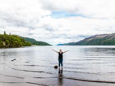 Young woman standing knee-deep in Loch Ness, surrounded by rolling green hills and rippling water