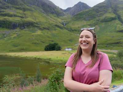 Smiling young woman in a t-shirt standing in Glen Coe valley, bathed in warm summer light