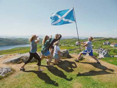Group of young travellers holding a Scottish flag on a sunny hilltop on the Isle of Lewis