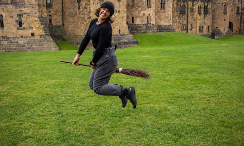 Young woman playfully pretending to fly a broomstick on the grassy grounds of Alnwick Castle