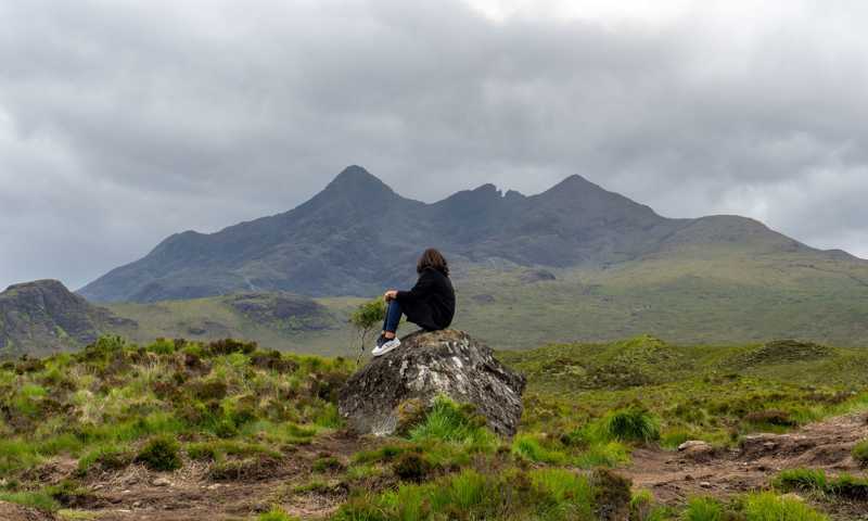 Young woman sitting on rock and enjoying views of rocky cuillin mountains on isle of skye